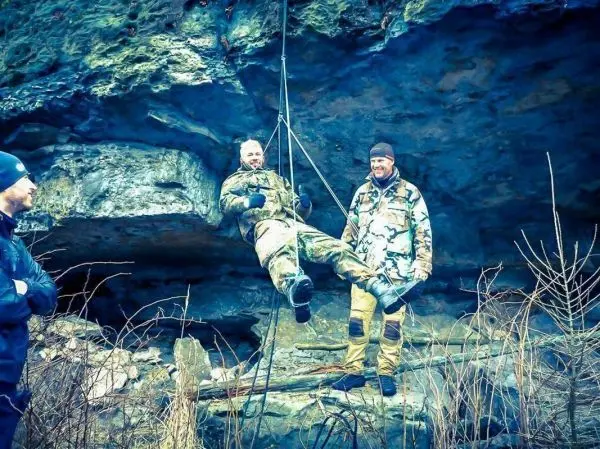 Zwei Personen stehen neben einem hängenden Mann, der an einem Seil befestigt ist