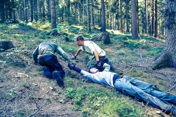 Drei Personen helfen einem am Boden liegenden Mann in einem Wald