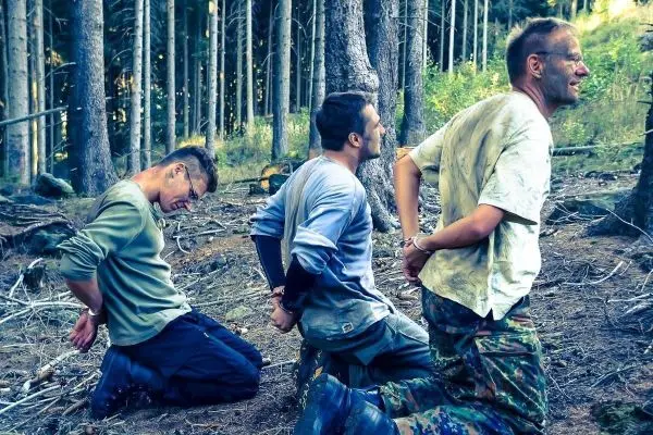 Drei Personen knien mit gefesselten Händen im Wald auf dem Boden