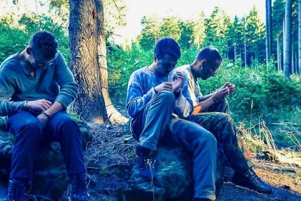 Drei Personen sitzen auf einem Stein im Wald, Hände gefesselt