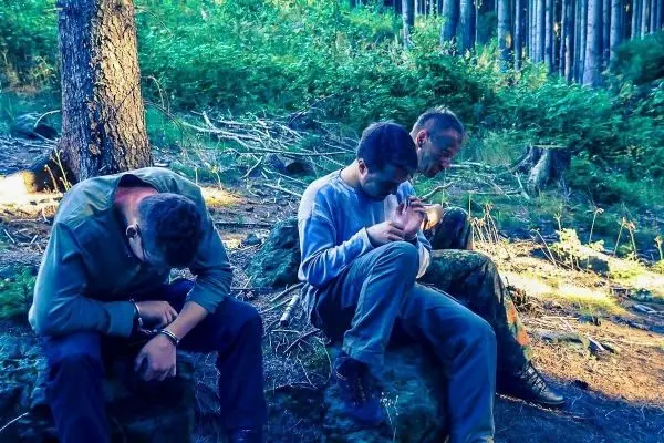 Drei Personen sitzen auf Steinen im Wald, Hände gefesselt, mit gesenkten Köpfen