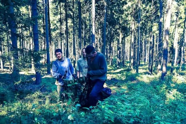 Drei Personen tragen Ausrüstung durch einen Wald mit hohen Bäumen und dichtem Unterholz