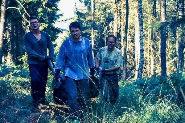 Drei Personen tragen eine große Tasche durch einen Wald mit hohen Bäumen