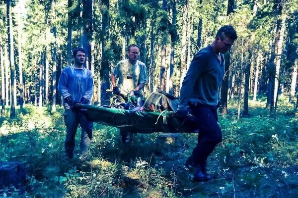 Drei Personen tragen eine improvisierte Trage durch einen Wald