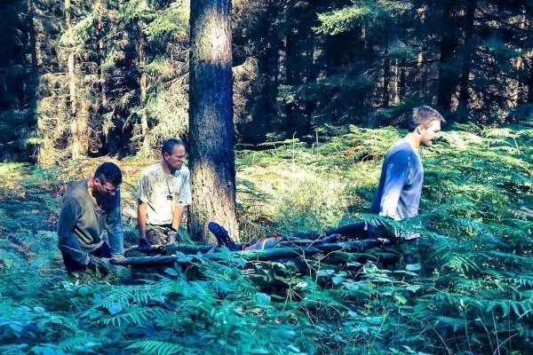 Drei Personen tragen eine improvisierte Trage durch einen Wald mit dichtem Unterholz
