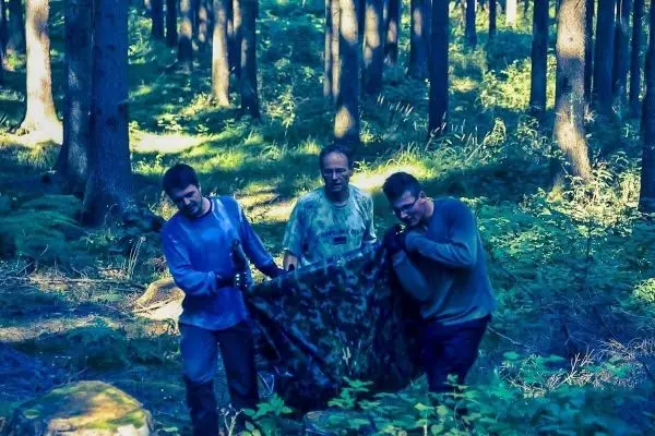 Drei Personen tragen eine Tarp-Notunterkunft durch den Wald