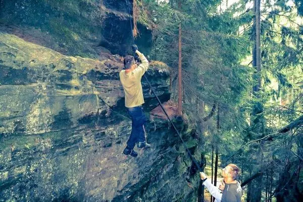 Eine Person klettert an einer Felswand, während eine andere sie mit einem Stock unterstützt