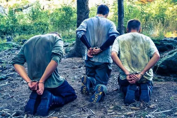 Es sind drei Personen in Handschellen, die auf dem Boden knien