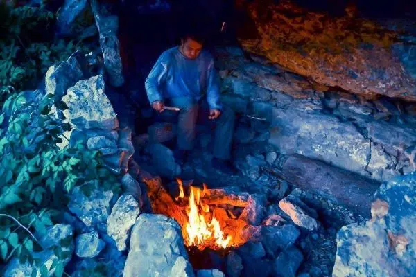 Kleines Bodenfeuer brennt zwischen Steinen, Person sitzt davor und hält Holzstücke