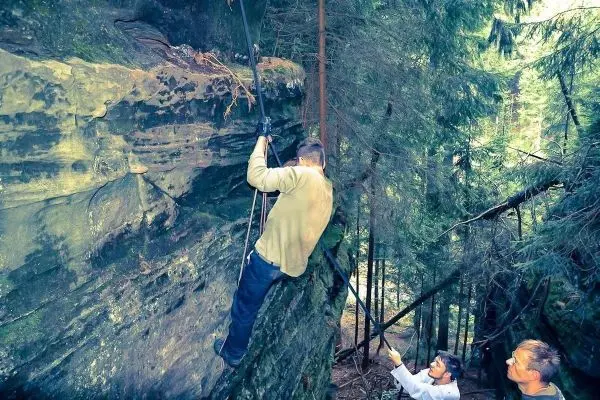 Mann klettert mit Seil an einer Felswand, zwei Personen sichern unten
