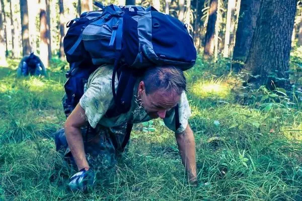 Mann krabbelt durch hohes Gras mit einem großen Rucksack auf dem Rücken