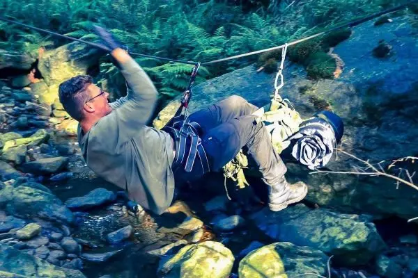 Mann überquert einen Bach an einer selbstgebauten Seilbrücke mit Ausrüstung