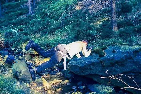 Person überquert einen kleinen Bach im Kriechgang, Ausrüstung liegt am Ufer