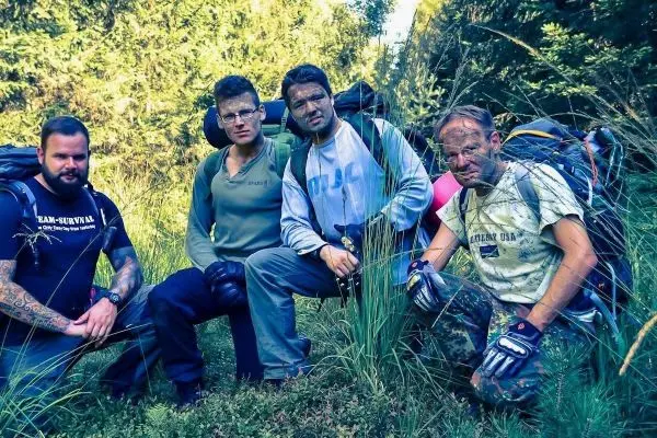 Vier Personen mit Rucksäcken knien im hohen Gras, Gesicht und Hände sind verschmutzt