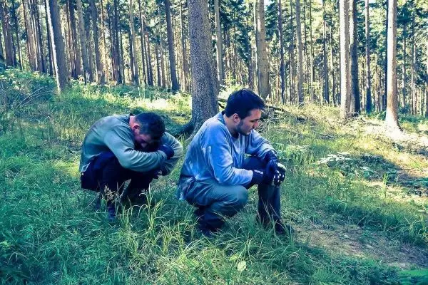 Zwei Personen hocken in einem Waldgebiet auf dem Boden zwischen Bäumen