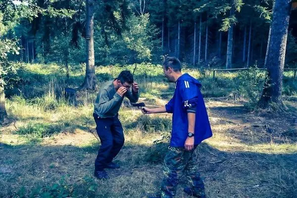 Zwei Personen in einem Wald, eine Person hält eine Waffe, die andere schützt sich