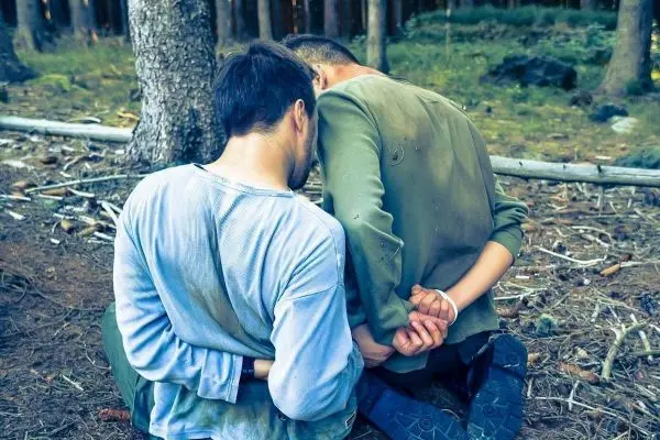 Zwei Personen mit gefesselten Händen knien im Wald auf dem Boden