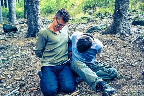 Zwei Personen sitzen auf dem Boden im Wald mit gefesselten Händen