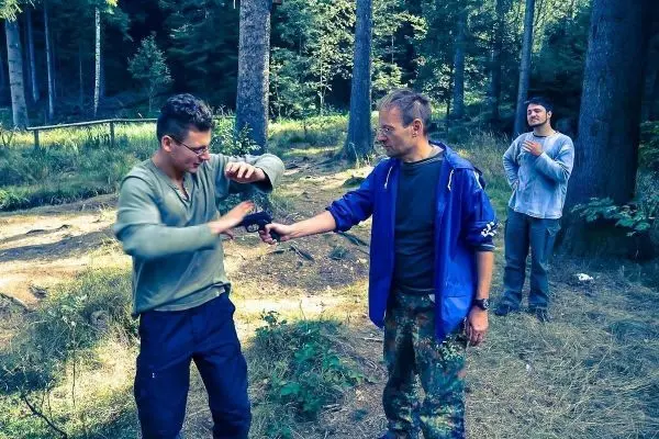Zwei Personen üben eine Selbstverteidigungstechnik im Wald, während eine dritte Person zuschaut