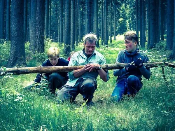 Drei Personen heben gemeinsam einen langen Stock im Wald