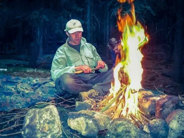 Mann sitzt vor einem Feuer und bearbeitet Holz mit einem Messer