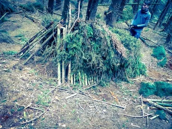 Person steht neben einer selbstgebauten Unterkunft aus Ästen und Zweigen