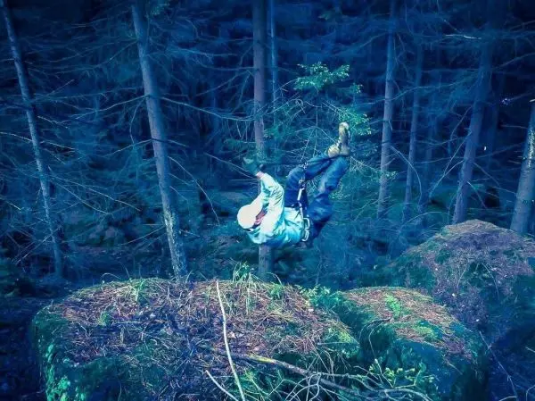 Person überquert mit Sicherheitsgurt eine selbstgebaute Seilquerung zwischen Bäumen