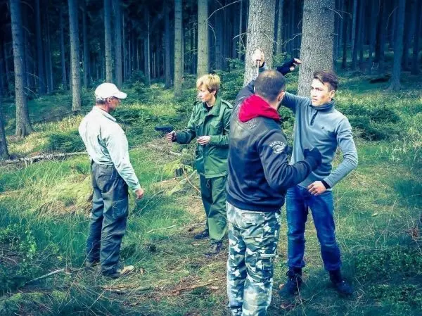 Vier Personen stehen im Wald, eine Person hält eine Waffe in der Hand