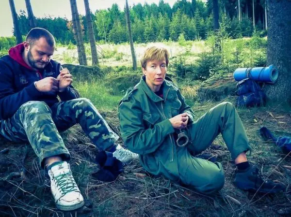 Zwei Personen sitzen im Wald, eine Person hält ein Messer und bearbeitet Holz