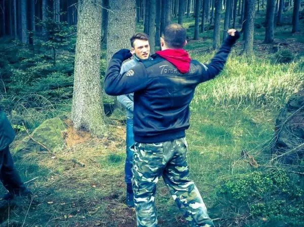 Zwei Personen stehen in einem Wald, eine Person hebt den Arm in einer gestikulierenden Bewegung