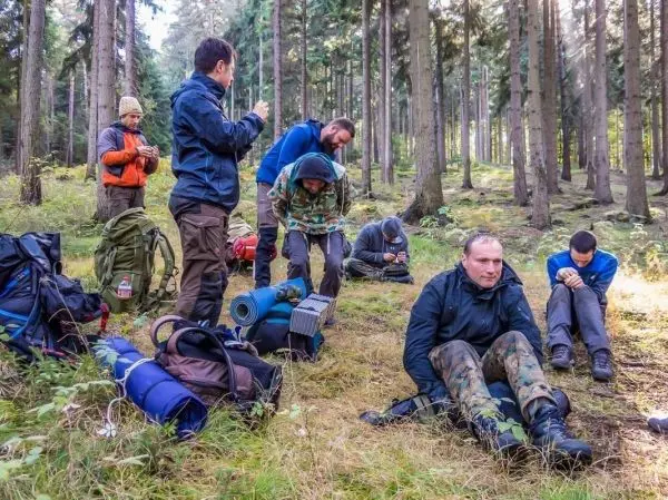 Gruppe sitzt und steht im Wald, Rucksäcke und Ausrüstung liegen auf dem Boden