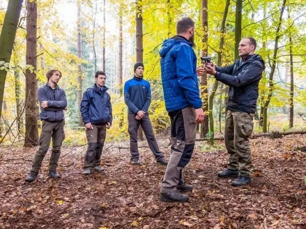 Mann demonstriert Ausrüstung an einem anderen Mann im Wald, umgeben von vier weiteren Personen
