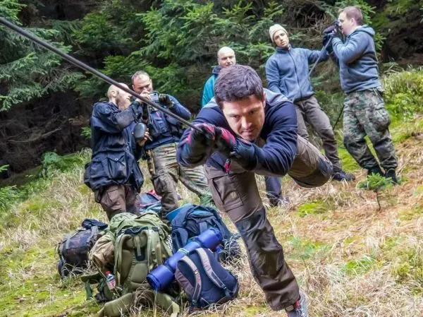 Mann überquert mit einem Seil eine unebene Fläche, Rucksäcke im Hintergrund