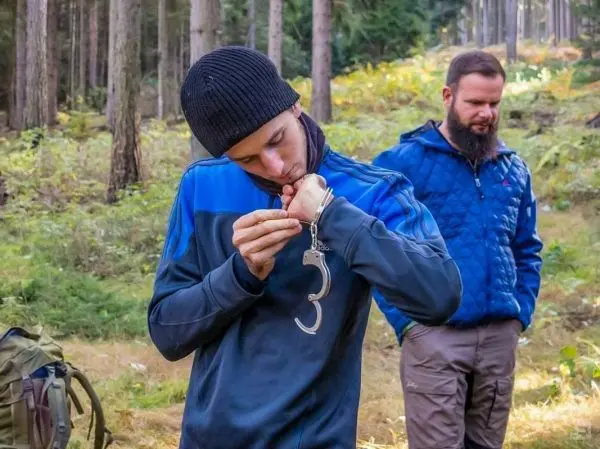 Mann untersucht Handschellen im Wald, ein weiterer Mann steht im Hintergrund