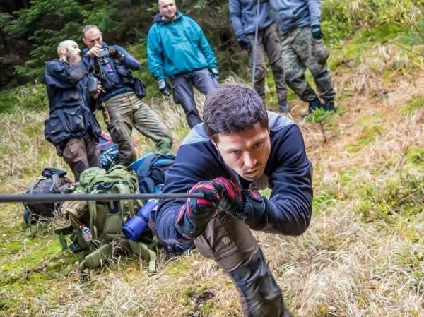 Mann zieht sich an einem Seil über unebenes Gelände, Rucksäcke im Hintergrund