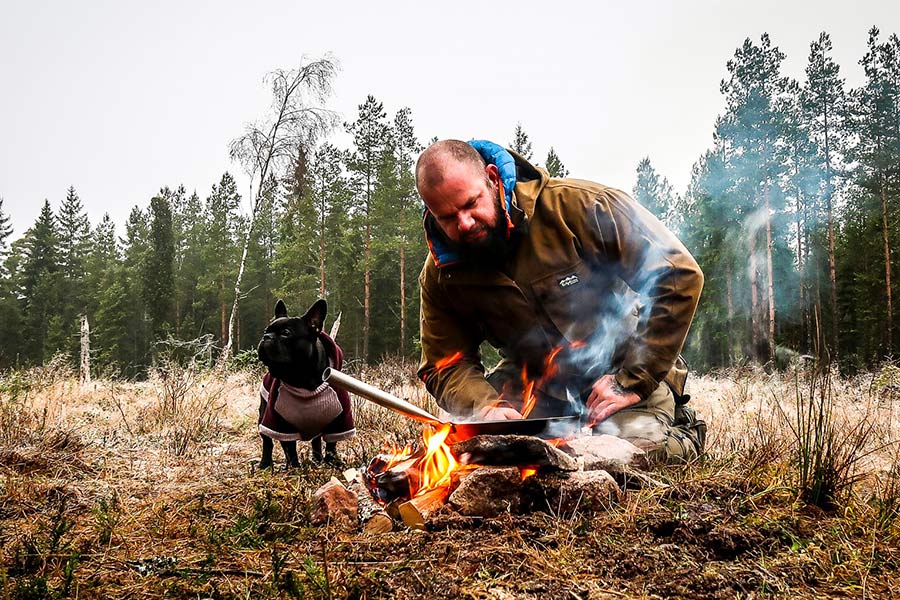 Ronny brät Speck in einer Pfanne über dem Lagerfeuer vor dem Wald