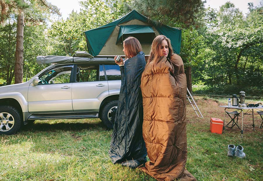 2 Frauen stehen in Schlafsäcken vor einem Auto