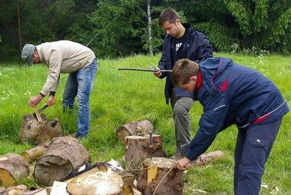 Drei Personen bearbeiten Holzstücke mit Messern und Äxten auf einer Wiese