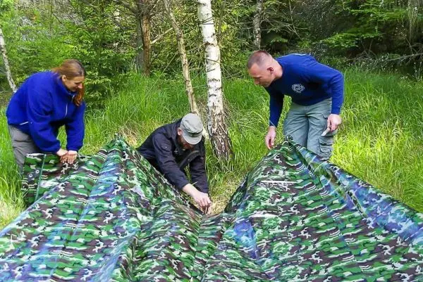 Drei Personen richten eine camouflagefarbene Plane im Gras aus