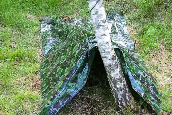 Eine selbstgebaute Unterkunft aus Tarnmaterial steht neben einem Baum