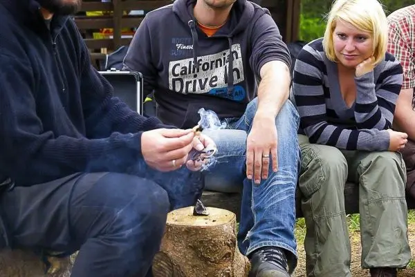 Kleinfeuer raucht auf einem Baumstumpf, während Personen aufmerksam zuschauen