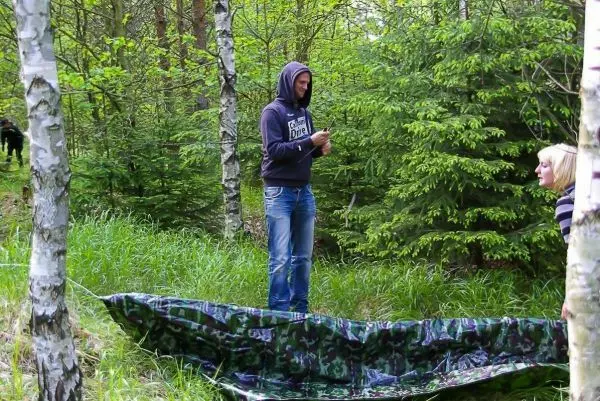 Mann mit Kapuze hält ein Seil neben einer Tarp-Notunterkunft im Wald