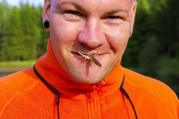 Mann mit orangefarbener Jacke hält eine Heuschrecke vor dem Mund