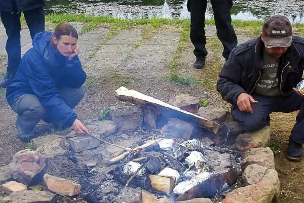 Person sitzt neben Feuerstelle und hält Stock über die Glut beim Kochen
