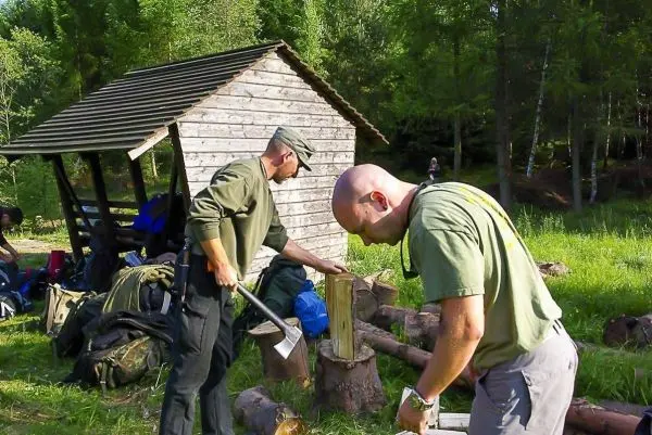 Zwei Personen bearbeiten Holz mit einer Axt und einem Messer neben einer Holzunterkunft