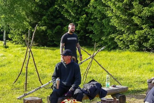 Zwei Personen sitzen auf einer Holzbank, umgeben von Bäumen und improvisierten Stangen
