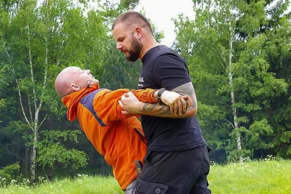 Zwei Personen üben eine körperliche Technik im Freien, umgeben von Bäumen