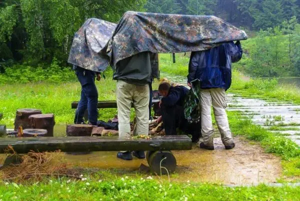 Gruppierung unter Tarp-Notunterkunft, Holzbank und Bodenfeuer in feuchter Umgebung