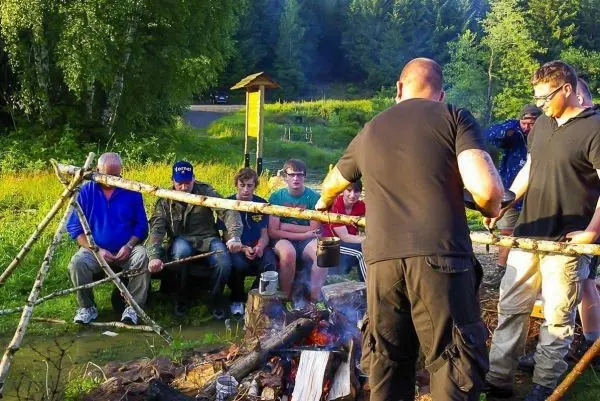 Kessel hängt über dem Feuer, während Teilnehmer am Lagerfeuer sitzen