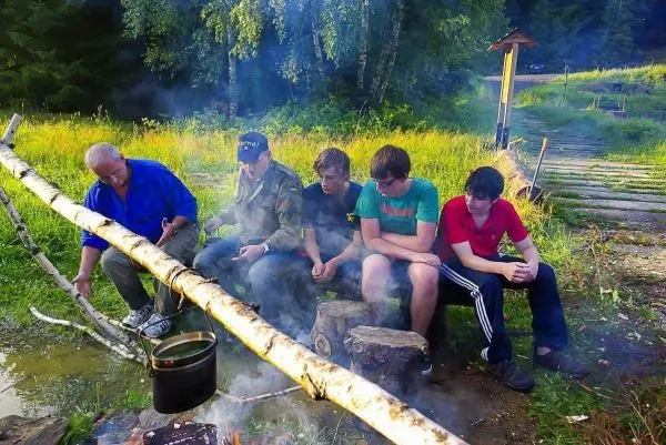 Kessel hängt über kleinem Feuer, Rauch steigt auf, Gruppe sitzt auf Baumstämmen